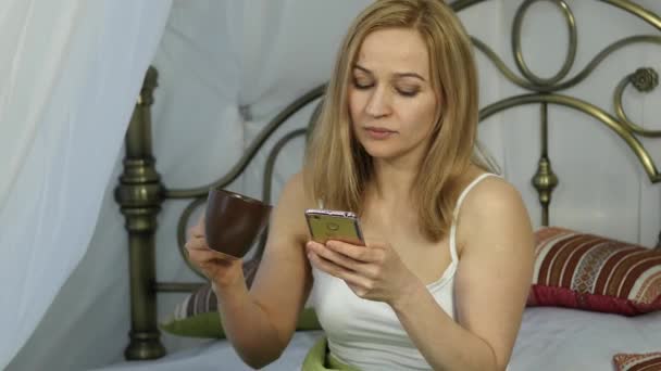 Veselá žena sedí v posteli tancovat a zpívat, pití kávy a chatování s přáteli na telefonu. 4k — Stock video