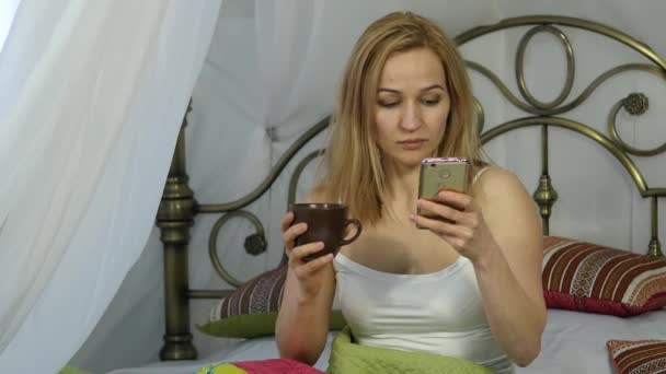 Femme gaie assise au lit dansant et chantant, buvant du café et bavardant avec des amis sur un téléphone. 4K — Video
