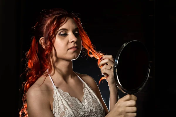 Молодая рыжая женщина держит круглое зеркало дома и исправляет свои волосы на темном фоне — стоковое фото