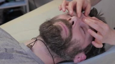 Adam yüz masajı yaptırıyor. Güzellik prosedürü, cilt yaşlanmasının önlenmesi. Kasmetolojistin ofisinde yatıyor. Alnında kırışıklıklar