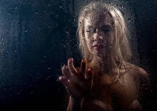 Beautyful blonde woman behind wet glass on a dark background, woman takes a shower — Stock Photo, Image