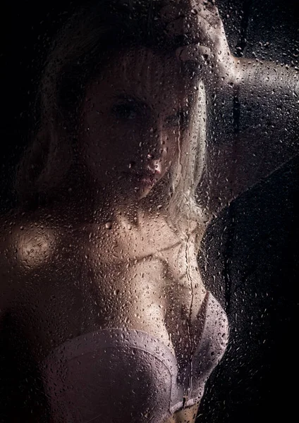 Schöne blonde Frau hinter nassem Glas auf dunklem Hintergrund, Frau duscht Stockbild