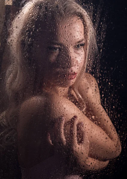 Vacker blond kvinna bakom vått glas på en mörk bakgrund, kvinna tar en dusch Royaltyfria Stockbilder