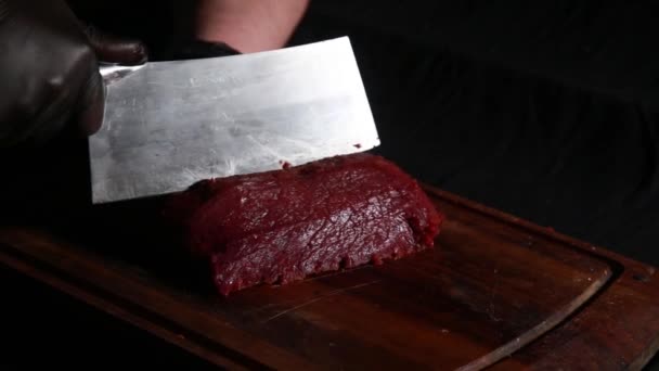 Chef snijdt rauw vlees met groot mes op een houten plank in het donker — Stockvideo