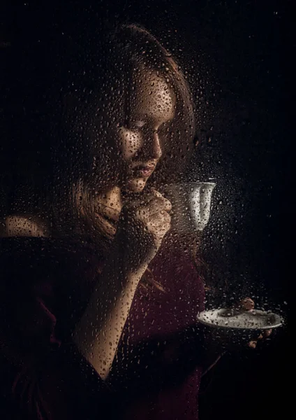 Nice young girl drinks coffee behind the wet glass, raindrops on glass — Stock Photo, Image