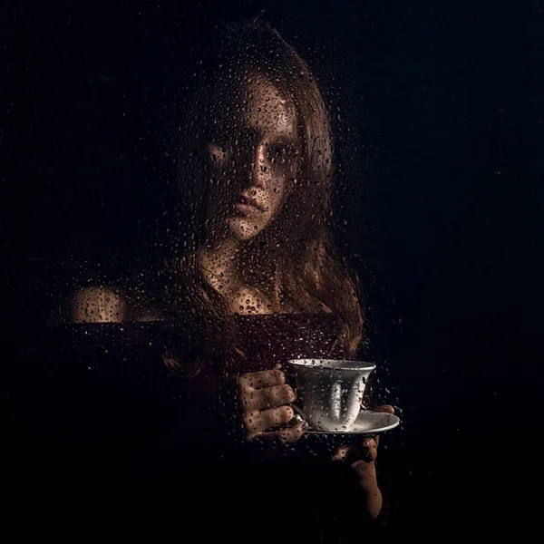 Belle jeune fille boit du café derrière le verre mouillé, gouttes de pluie sur le verre — Photo