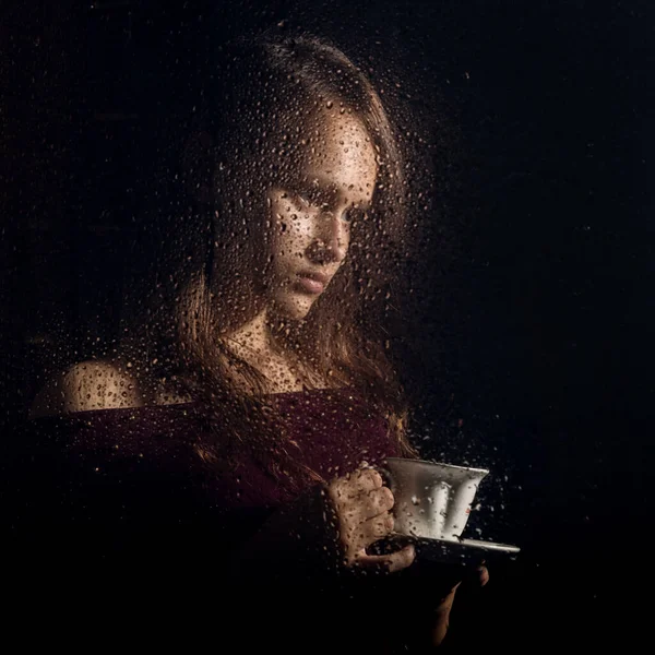 Belle jeune fille boit du café derrière le verre mouillé, gouttes de pluie sur le verre — Photo