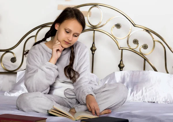 young student girl reads a book while lying on a bed doing homework