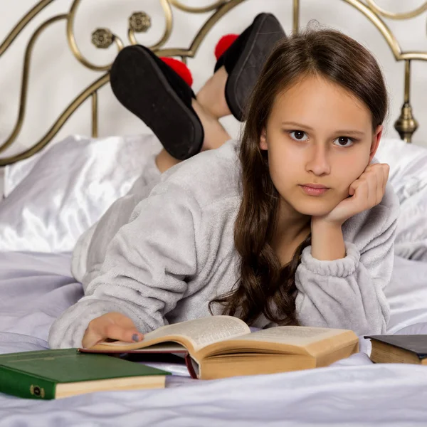 Junge Studentin liest ein Buch, während sie auf einem Bett liegt und Hausaufgaben macht — Stockfoto
