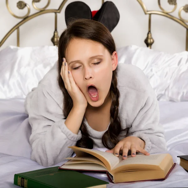 Sad young student girl reads a book while lying on a bed doing homework — Stock Photo, Image