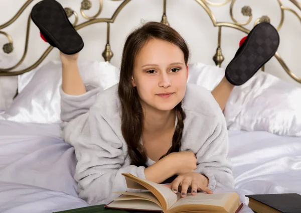 Mladá studentka čte knihu, zatímco leží na posteli a dělá domácí úkoly — Stock fotografie
