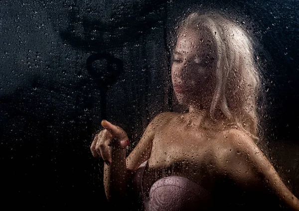 Beautyful blonde woman behind wet glass on a dark background, woman takes a shower — Stock Photo, Image