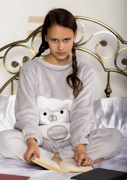 Junge Studentin liest ein Buch, während sie auf einem Bett liegt und Hausaufgaben macht — Stockfoto