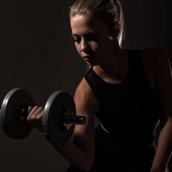 Fitness menina sexy com halteres em um fundo escuro. Atleta fazendo exercícios no ginásio — Fotografia de Stock