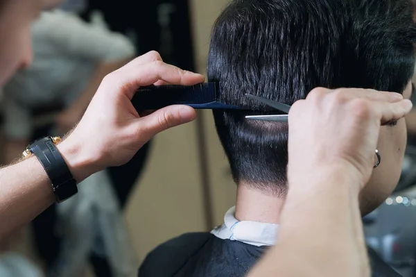 Cabeleireiro mestre cortando um jovem atalho na barbearia. cliente de cabelos escuros em capa brilhante sentado na cadeira na barbearia . — Fotografia de Stock