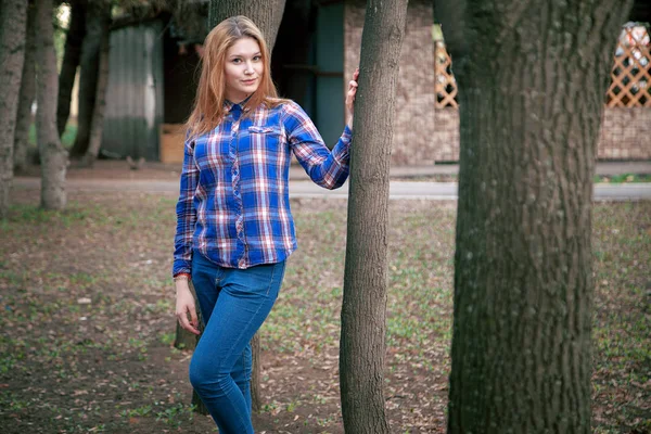 Retrato de uma menina bonita. sorrindo, posando na câmera. numa camisa azul numa jaula. No contexto do parque de outono — Fotografia de Stock