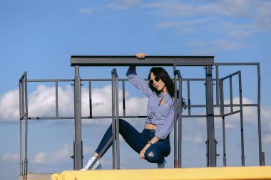 Güneş gözlüğü ve yırtık kot pantolon ve çizgili bir gömlek ile güzel bir kız. Büyük çelik yapılar arka planı. Sanatsal fotoğraf. Güzellik ve moda, stil