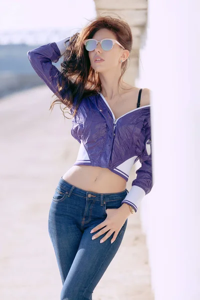 Mujer joven de moda en gafas de sol, cortavientos cortos lila y jeans. Se encuentra sobre una estructura de hormigón, hombro desnudo. Moda femenina. Modo de vida urbano — Foto de Stock