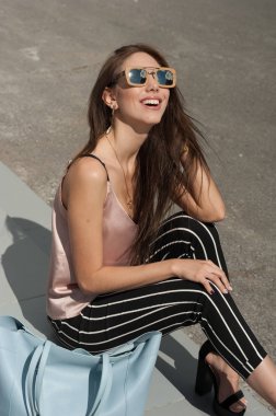 Güneş gözlüğü, ahşap bir çerçeve içinde giyen genç güzel bir moda mutlu Bayan açık portresi. Modeli giyer şık giysiler, bej bir bluz, beyaz bir şerit içinde siyah pantolon, bir çanta dolusu ışık bl