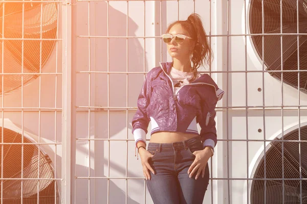 Модная молодая женщина в солнечных очках, сиреневой короткой ветровке и джинсах. на фоне больших кондиционеров воздуха. Женская мода. Городской образ жизни — стоковое фото