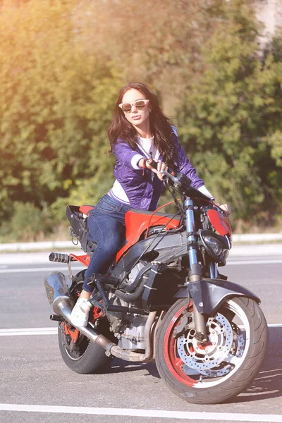 Fashionable young woman with sunglasses, lilac short windbreaker and jeans with a beautiful body on a red sports bike — Stock Photo, Image