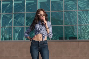Genç, güzel esmer kadın bir gözleme boynuz pembe bir dondurma yemek. İyi bir ruh halinde o siyah gözlük, bir gömlek bir beyaz ve mavi şerit ve koyu mavi jeans ile gözlük takıyor