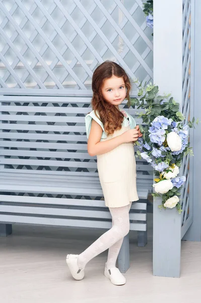 Yedi yıllık kestane rengi saçlı küçük bir kız yanındaki bir ahşap Bank durgunluk ve gri-mavi bide fular çiçekli parlak giysilerle gülümseyen bukleler. Bir tatil kavramı. — Stok fotoğraf