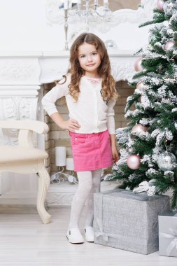 Yedi bir Noel ağacı ve hediyeler, kestane kıvırcık saçlı tatlı bir küçük kız pembe bir etek, beyaz bir kazak, kamera için poz duruyor. Bir tatil, Noel kavramı