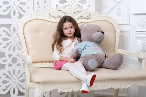 Yedi kestane kıvırcık saçlı küçük bir kız bir kucaklama zemin ışık bir ekranın karşı bir oyuncak ayı ile yumuşak mobilyalar oturur. Aşk ve kutlama kavramı — Stok fotoğraf
