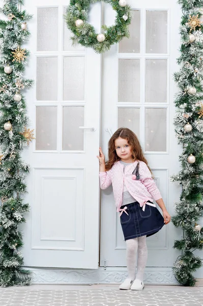 Yedi yıllık kestane renginde saçlı tatlı bir küçük kız, Katlanır Kapılar arka plan üzerinde duruyor bir Noel tarzda bir mavi etek, pembe bir ceket ve kamera için poz dekore edilmiş — Stok fotoğraf