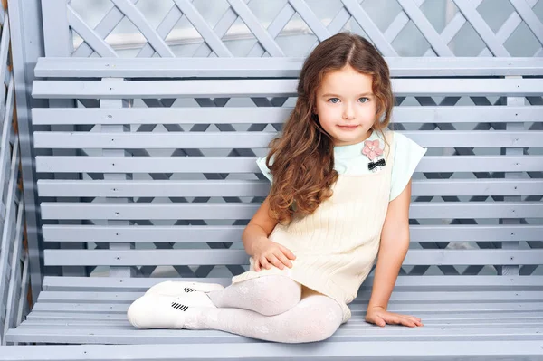 Yedi kestane rengi saçlı küçük bir kız bir ahşap Bank bir gri-mavi renkte parlak giysiler ve bukleler ile gülümseyen oturur. Bir tatil kavramı — Stok fotoğraf