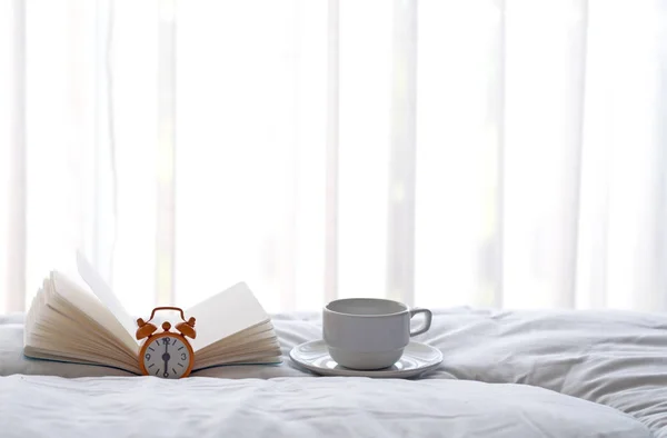 Будильник на кровати с кофе и книга утром с солнечным светом — стоковое фото