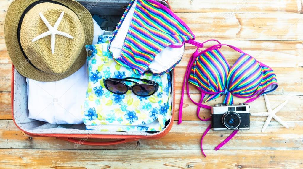 Open the suitcase with tourist things: women's hat, swimsuit, camera, denim shorts, dresses, sunglasses, perfumes, tablet on wooden background