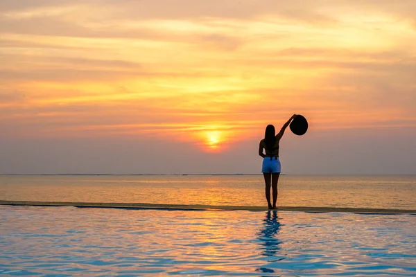 Mulher segurando chapéu relaxante na piscina ao pôr do sol e crepúsculo — Fotografia de Stock