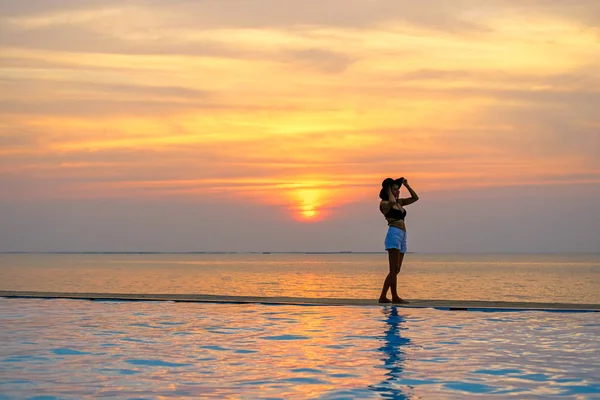 Mujer sosteniendo sombrero relajante en la piscina en el atardecer y crepúsculo —  Fotos de Stock