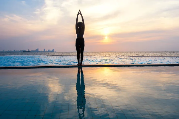 剪影年轻女人上练瑜伽的游泳池和海滩日落时分. — 图库照片