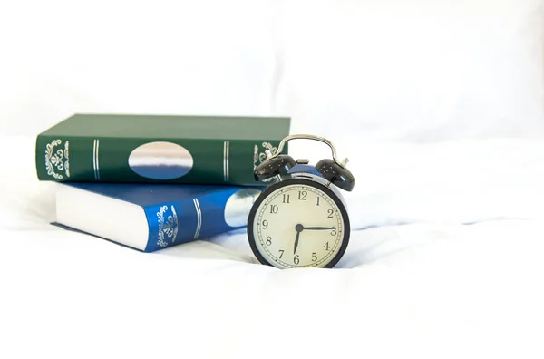 Relógio despertador com livro na cama de manhã com luz do sol — Fotografia de Stock