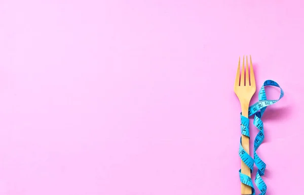 青の測定テープはカラフルなピンクの背景に横たわっている木製のフォークを包んだ。ダイエットと健康の概念. — ストック写真