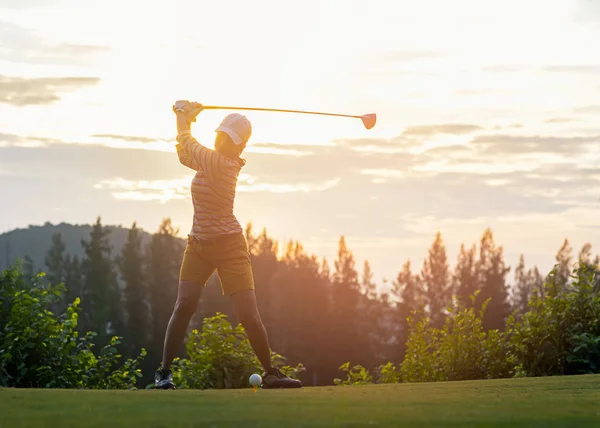 Asian woman golf player doing golf swing tee off on the green sunset evening time, she presumably does exercise.  Healthy and Lifestyle Concept.
