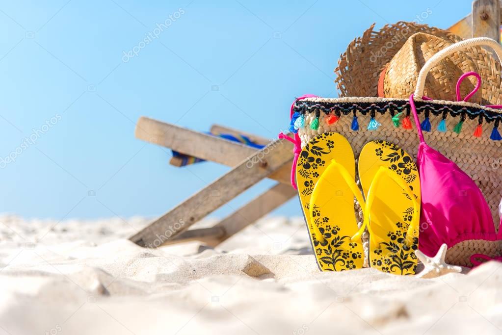 Summer Travel. Bikini and Flip-flops ,hat, fish star and bag near beach chair on sandy beach against blue sea and sky background, copy space.  Summer vacation concept