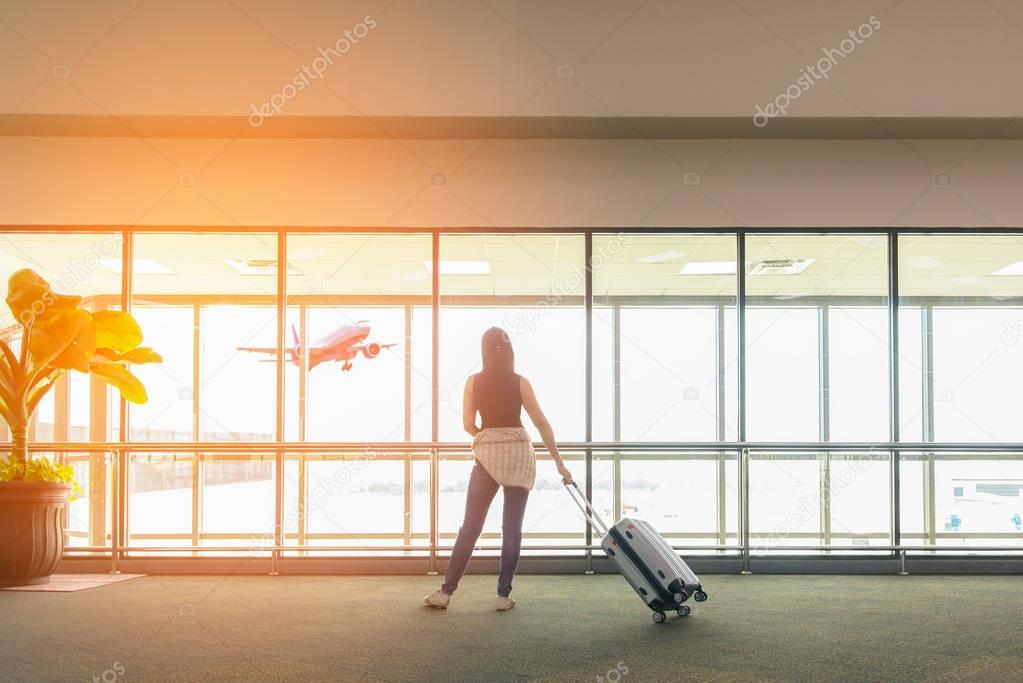 Traveler woman standing and hand holding luggage bag see the airplane at the airport glass window.  Tourist waiting in hall airplane departure. Travel Concept .