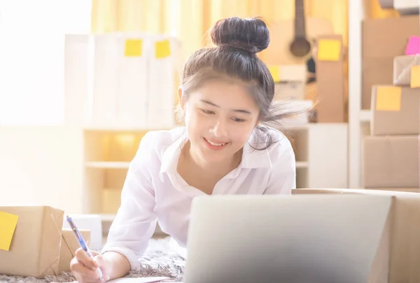 Asian smiling young women writing order from customer online for preparing package product.  Lifestyle for working at home.
