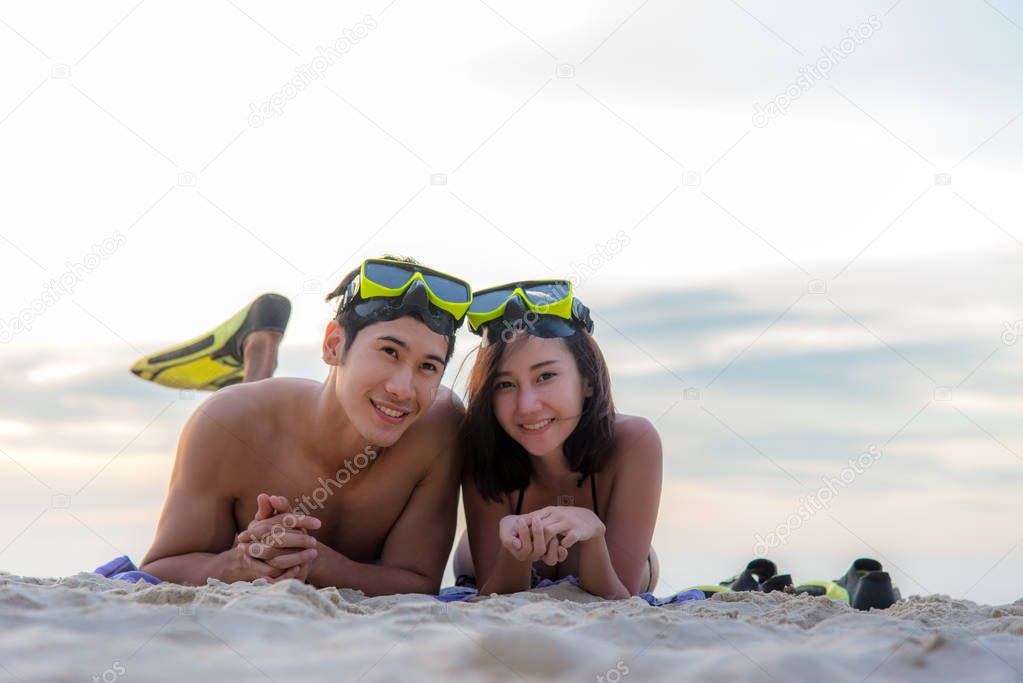 Summer Vacations. Beach travel couple having fun snorkeling. Asian smiling Couple lying  and enjoy on summer beach sand with snorkel equipment looking to side after swimming with fins and mask on vacation. Travel Concept