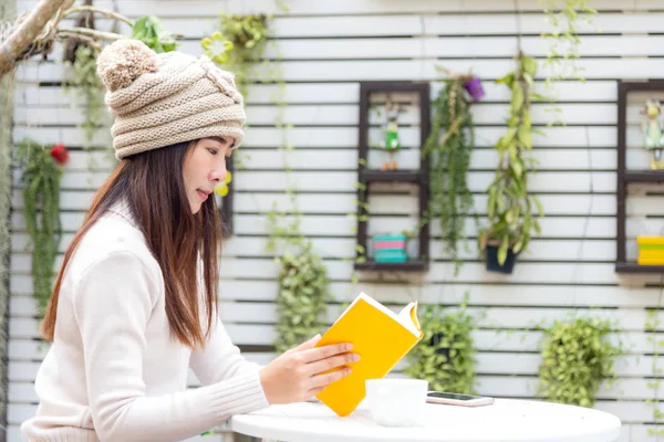 セーターを着たアジアの若い女性は 冬の季節に家の庭で熱いコーヒー スマートフォンで座って本を読んでいます ライフスタイルコンセプト — ストック写真