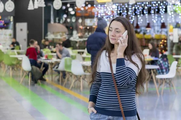 Jovem mulher andando no centro comercial e falando em um smartphone branco — Fotografia de Stock