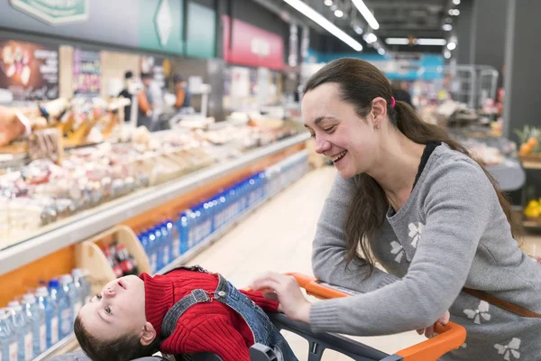 Una joven con un hijo pequeño en un supermercado. El chico se sienta en un carrito de bebé y se ríe — Foto de Stock
