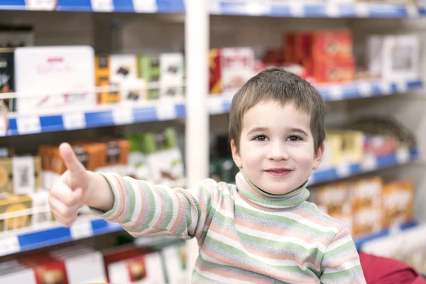 Menino feliz 4 anos em um supermercado no fundo de prateleiras com chocolate. O rapaz mostrou que está tudo bem. — Fotografia de Stock