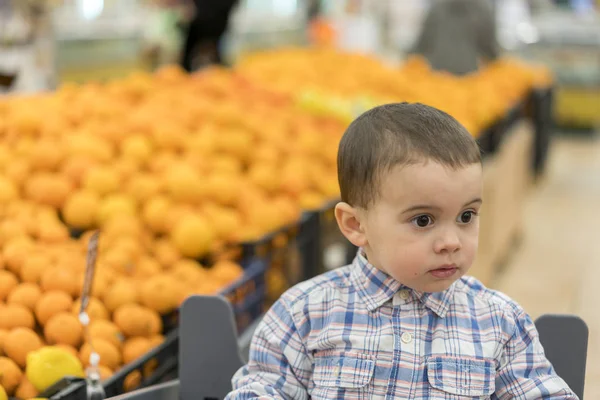 Симпатичный мальчик, одетый в клетчатую рубашку в супермаркете на тележке. На фоне апелляций — стоковое фото