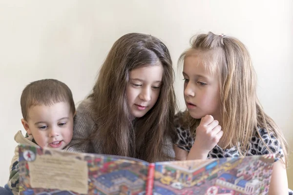 Tres niños pequeños leen un libro grande. Dos hermanas y un hermano. Fondo claro. Aspecto europeo —  Fotos de Stock