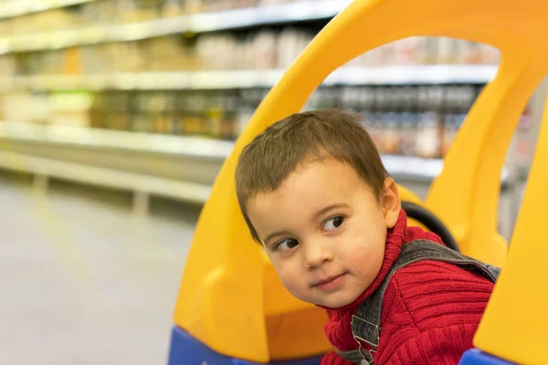 Niño pequeño mira desde un carrito de compras para niños en un supermercado en el fondo de los estantes con comida — Foto de Stock
